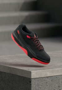 Μαύρο αθλητικό παπούτσι με κόκκινες λεπτομέρειες, διαθέτοντας λείο ανώτατο υλικό, υφασμένο μαύρο δίχτυ με υφή και κορδόνια με σχέδιο, τοποθετημένο στον αέρα πάνω από σκυρόδεμα.