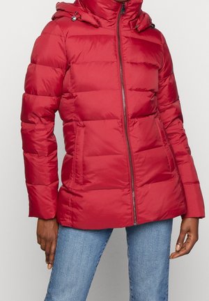 Personne portant une doudoune matelassée rouge avec une fermeture éclair frontale et une capuche, associée à un jean bleu clair, debout devant un fond uni.