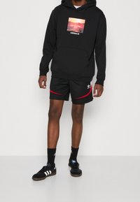 Felpa nera con cappuccio e stampa paesaggistica, pantaloni corti neri con accenti rossi, sneakers nere con strisce bianche e calzini neri.