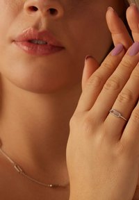 Zilveren ring met een verbonden ovaal ontwerp, versierd met kleine heldere steentjes. De hand van het model raakt voorzichtig haar gezicht aan met milde roze nagellak.