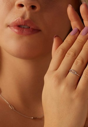 Silver ring with linked oval design, adorned with small clear stones. Model's hand gently touches face with mild pink nail polish.