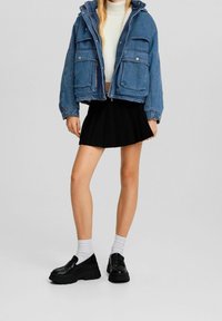 Femme portant une veste en jean bleu, un crop top blanc à col roulé, une jupe plissée noire, des chaussettes blanches et des mocassins à plateforme noirs, se tenant sur un fond gris.