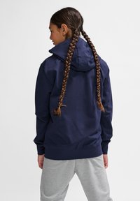Marineblauer Hoodie mit einer doppellagigen Kapuze, gerippten Bündchen und Saum. Trägt graue Jogginghose, die Haare in zwei Zöpfen gestylt.