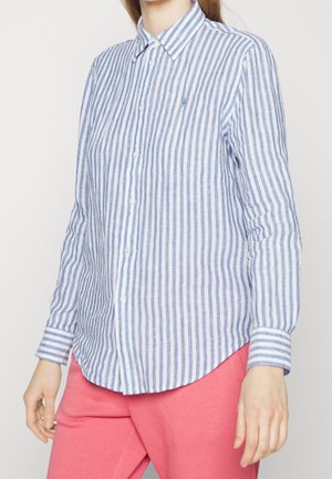 Kvinde iført en blå og hvid lodret striped skjorte med krave og lyserøde bukser mod en ensfarvet baggrund.