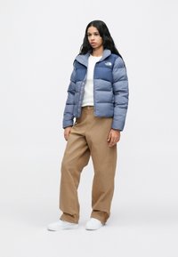 Egy nő kétrétegű kék pufferszövet kabátot, fehér pólót, széles bézs nadrágot és fehér sportcipőt visel, háttérben egy sima fehér fal.