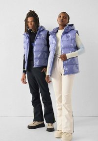 Twee personen dragen glanzende paarse donsbodywarmers met capuchons, zwarte en crèmekleurige broeken en beige laarzen. De achtergrond is neutraal.