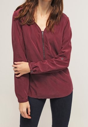 Kobieta ubrana w ciemnoczerwoną bluzkę z długim rękawem i zamkiem z przodu, stoi z jedną ręką skrzyżowaną na drugą, w ciemnych spodniach.