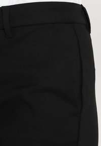 Close-up van de taille van zwarte op maat gemaakte broeken met riemlussen en een zijzaknaad op een witte achtergrond.