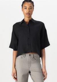 Camicia nera a maniche corte, con colletto e bottoni, dal taglio cropped e dalla texture liscia, abbinata a pantaloni grigi chiari aderenti.