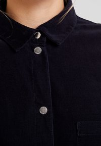 Primo piano del colletto di una camicia in corduroy blu scuro con tre bottoni a pressione argentati visibili e vista parziale della tasca sul lato destro.