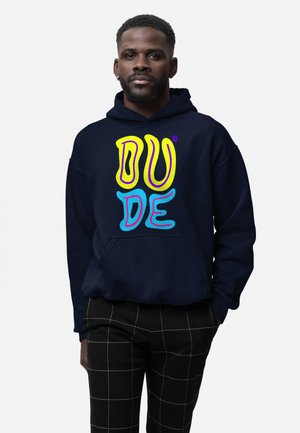 Hombre con sudadera con capucha azul marino con texto colorido "DUDE" y pantalones negros con patrón de cuadrícula blanca, de pie contra un fondo blanco.