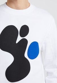 Sweat-shirt blanc avec de grandes formes abstraites en noir et bleu. Texture lisse, coupe à col rond, avec détails de couture sur les manches.