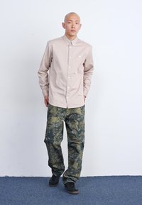 Světle béžová košile s dlouhým rukávem a límečkem, s logem na hrudi; kombinována s volnými kalhotami se zeleným camouflage vzorem a černými botami.