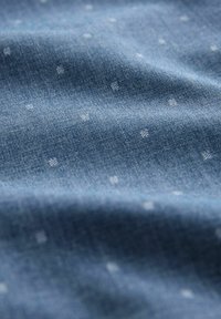 Tissu en denim bleu clair avec une surface texturée subtile, présentant de petits pois blancs régulièrement espacés.