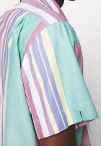 Uomo che indossa una camicia a maniche corte con righe verticali in verde pastello, rosa, blu, giallo e bianco su uno sfondo neutro.