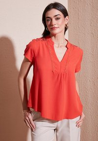 Blusa rossa a maniche corte con scollatura a V, caratterizzata da cuciture decorative lungo il collo e leggera arricciatura per una vestibilità comoda.