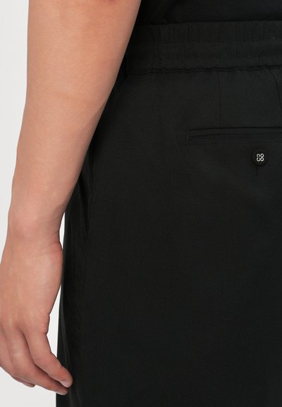 Pantalons en tissu noir avec une taille élastique, des poches latérales et un petit logo brodé sur la poche arrière. Matériau texturé.