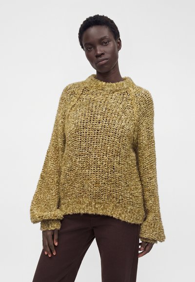 Femme aux cheveux courts et bouclés portant un pull ample texturé jaune moutarde et un pantalon marron foncé sur un fond blanc uni.