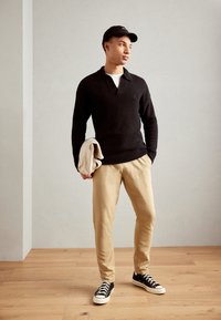 Black textured knit pullover, beige tapered pants, and black sneakers. Holding a beige garment. Simple design against a light background.