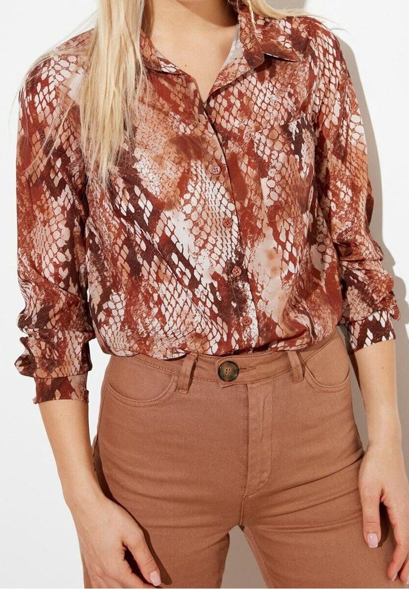 Femme portant une chemise à manches longues à imprimé serpent marron et beige, boutonnée et rentrée dans un pantalon taille haute marron.