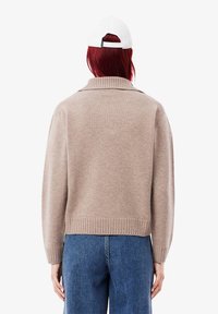Beiger gestrickter Pullover mit geripptem Saum und Kragen. Verfügt über überschnittene Schultern und lange Ärmel. Getragen mit blauen Jeans und einer weißen Kappe.