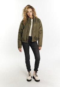 Giubbotto imbottito verde oliva con bottoni a scatto, abbinato a una maglietta beige a righe, jeans skinny neri e stivali bianchi alla caviglia con suole nere.