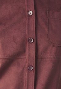 Bordowa koszula zamszowa o gładkiej strukturze, zapinana na cztery dopasowane guziki z przodu, z pojedynczą kieszeni�ą na piersi.