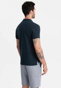 Tummansininen puuvillapolo, jossa on lyhyet hihat, sivuhalkiot ja selkeä muotoilu, yhdistettynä vaaleanharmaisiin shortseihin.