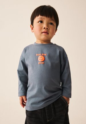 Niño pequeño con cabello corto y oscuro que lleva una camiseta azul de manga larga con una cara sonriente naranja y texto "Best Day Ever".