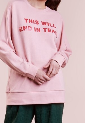 Osoba nosząca różową bluzę z czerwonym napisem "THIS WILL END IN TEARS" oraz ciemnozielone teksturowane spodnie.