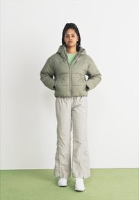 Giacca verde oliva imbottita con chiusura con zip, abbinata a pantaloni grigi chiari. Il modello è in piedi su un pavimento verde contro uno sfondo bianco.