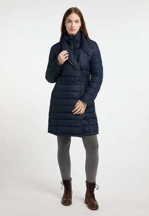 Marineblauwe pufferjas met een sjalkraag, een ceintuur in de taille en een horizontaal quiltpatroon, gecombineerd met grijze skinny jeans en bruine veterschoenen.
