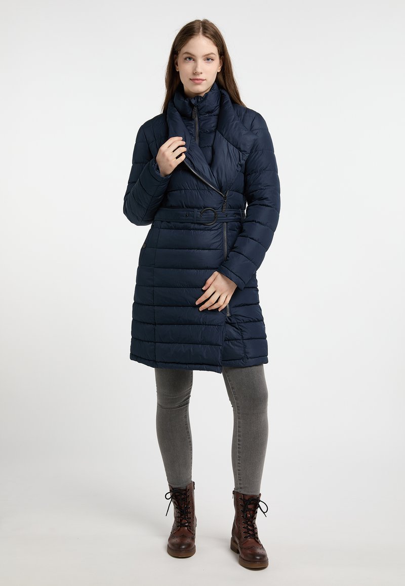 Geacă puffer bleu marin cu guler tip șal, talie cu curea și model matlasat orizontal, asortată cu blugi skinny gri și botine maro cu șireturi.