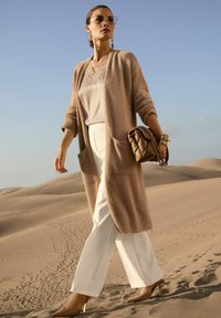 Frau in beigem Cardigan, weißen Hosen und hohen Schuhen hält eine braune, gesteppte Handtasche und steht auf Sanddünen unter einem klaren blauen Himmel.