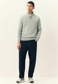 Man staat en draagt een lichtgrijze zip-necksweater, donkere jeans en grijze sneakers, met één hand in de zak tegen een effen lichte achtergrond.