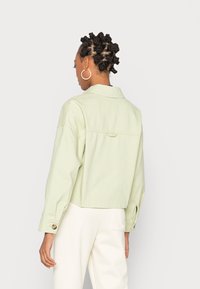 Svetle zelená zkrácená bunda z bavlny, s límečkem, knoflíkovými manžetami a detailem na zadní části. Nositá přes bílé kalhoty.