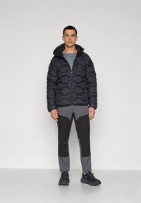 Giacca imbottita nera con design trapuntato e cappuccio, abbinata a pantaloni sportivi grigio e nero con motivo a fantasia. Indossata con scarpe da ginnastica scure.