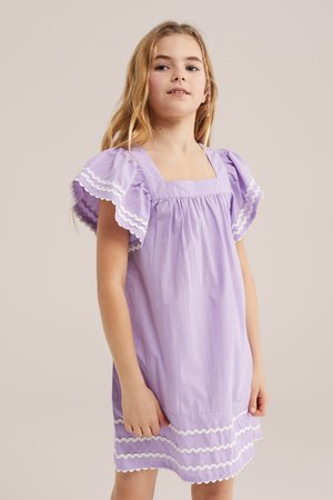 Fată cu păr lung și blond, îmbrăcată într-o rochie mov deschis cu mâneci bufante și margini albe, stând în fața unui fundal simplu.