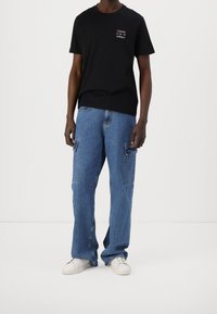 Osoba ubrana w czarną koszulkę Tommy Hilfiger, niebieskie dżinsy cargo i białe trampki, stojąca na tle jednolitego białego tła.