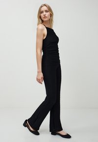 Jeune femme en haut sans manches noir et pantalon texturé assorti, marchant, portant des chaussures à petit talon noires, fond neutre.
