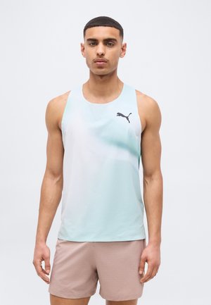 Bărbat tânăr purtând un tricou sport Puma fără mâneci, de culoare albastru deschis, și pantaloni scurți maro deschis, stând cu fața înainte pe un fundal simplu.