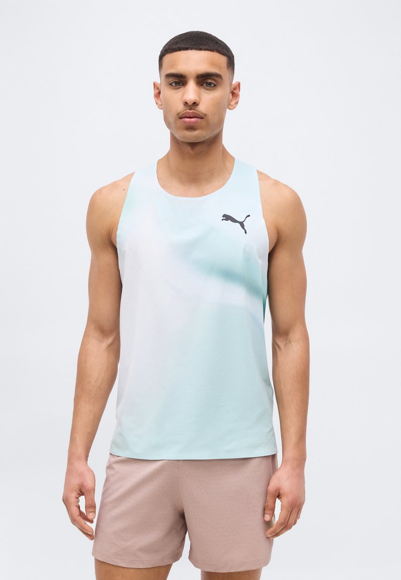 Νεαρός άνδρας που φοράει ανοιχτό μπλε αμάνικο αθλητικό μπλουζάκι Puma και ανοιχτό καφέ σορτς, στέκεται με το σώμα προς τα εμπρός μπροστά σε απλό φόντο.