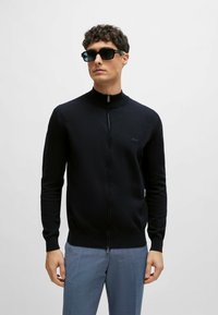 Felpa nera con zip e colletto alto, realizzata in materiale morbido, con logo ricamato sul petto. Abbinata a pantaloni leggermente blu con texture.