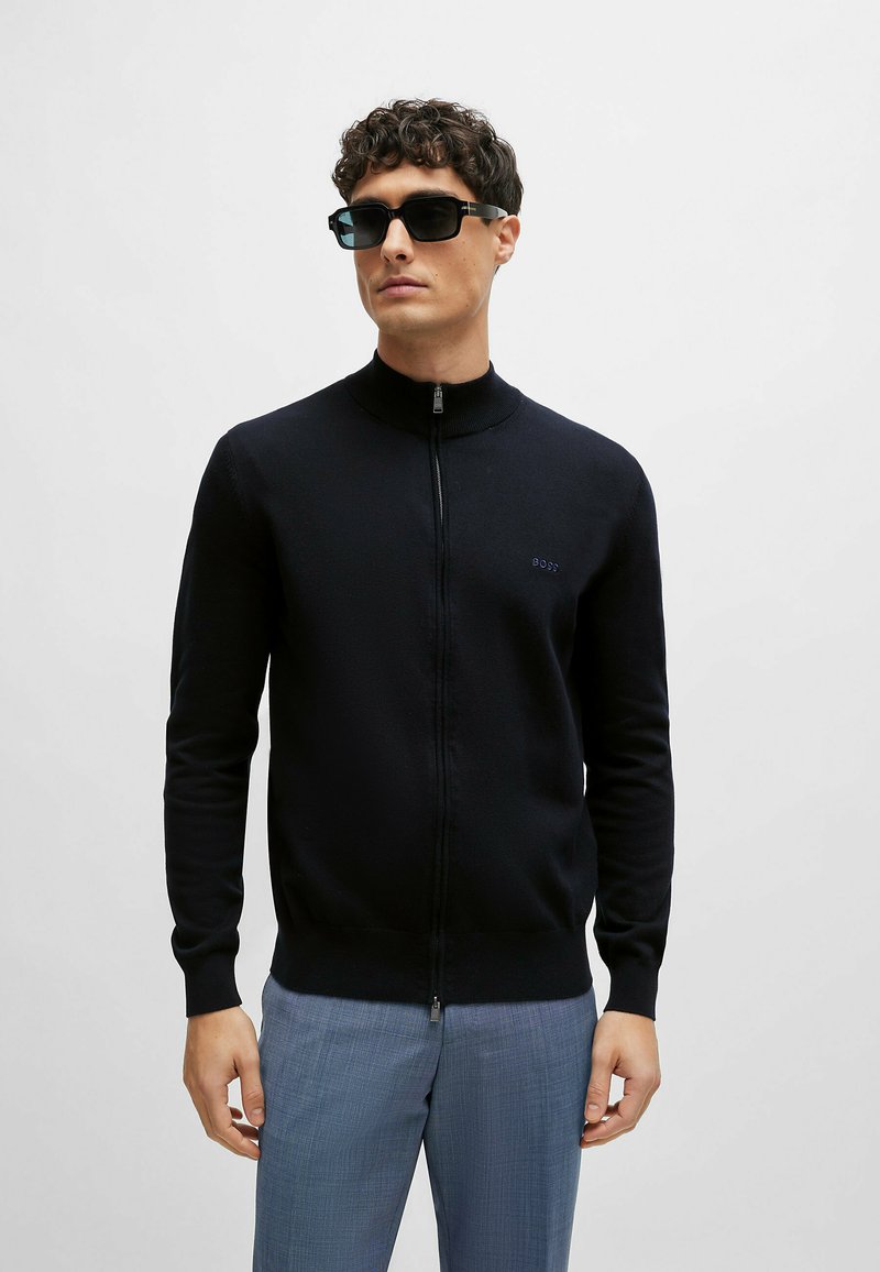 Felpa nera con zip e colletto alto, realizzata in materiale morbido, con logo ricamato sul petto. Abbinata a pantaloni leggermente blu con texture.