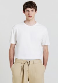 Giovane uomo con capelli corti castani che indossa una camicia bianca a maniche corte con una texture e pantaloni beige con una cintura in tessuto, mani in tasca, sfondo neutro.