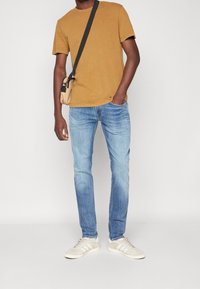 Brun t-shirt med kort ärm, ljusblå jeans och krämfärgade sneakers. Modellen håller också en liten beige axelväska. Enkelt, avslappnat set.