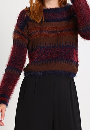 Femme portant un pull rayé duveteux bleu marine, bordeaux et marron à manches longues, associé à un pantalon noir ample.