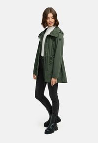 Zielona kurtka outdoorowa z kapturem, regulowanym sznurkiem w talii, zapięciem na zamek błyskawiczny i napy oraz czarnym logiem. Nosi ją wierzchnią warstwą na białym swetrze i czarnych dżinsach.