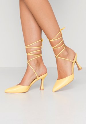 Żółte wysokie klapki na obcasie z ostrym noskiem, gładką skórzaną powierzchnią oraz cienkimi paskami wokół kostek, wiązanymi na łydkach.