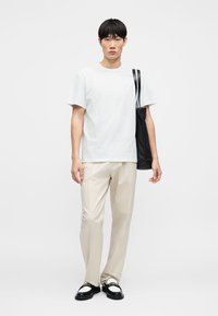 Jeune homme portant un t-shirt blanc, un pantalon beige, des mocassins noir et blanc, et portant un sac à bandoulière noir sur un fond uni.